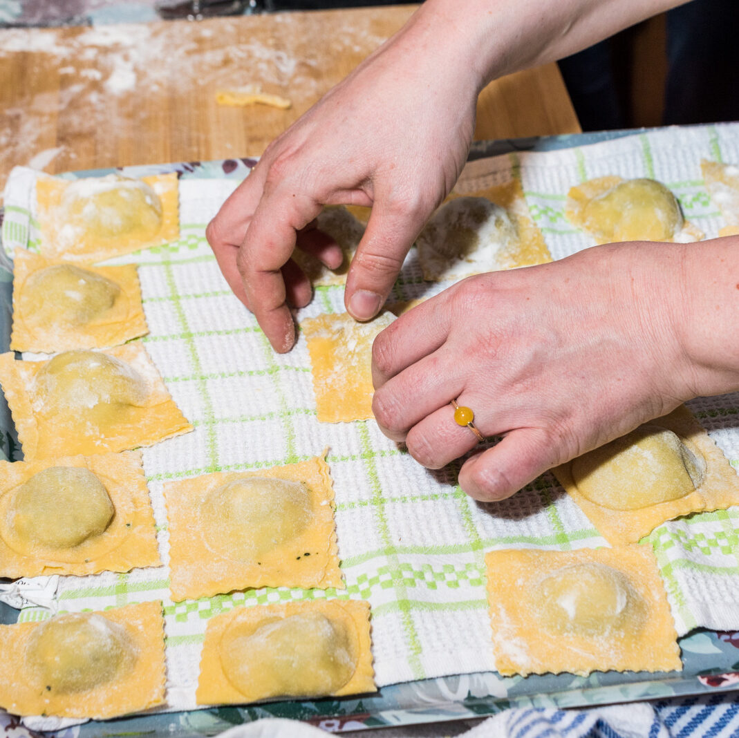 Close-up photo of hands touching raw, freshly-made ravioli surrounded by more ravioli in rows on a dishcloth.