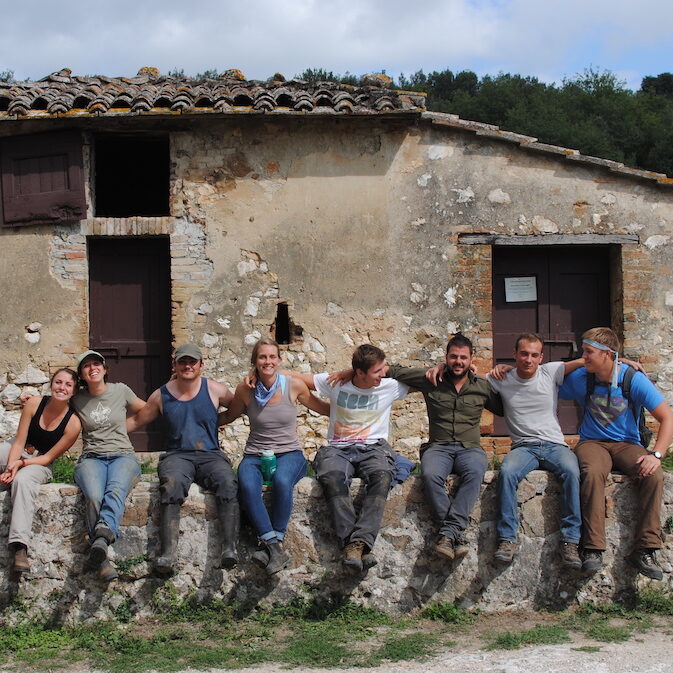 A group of interns sitting arm-in-arm along an old stone wall. They are in work clothes and an old stone building is behind them.