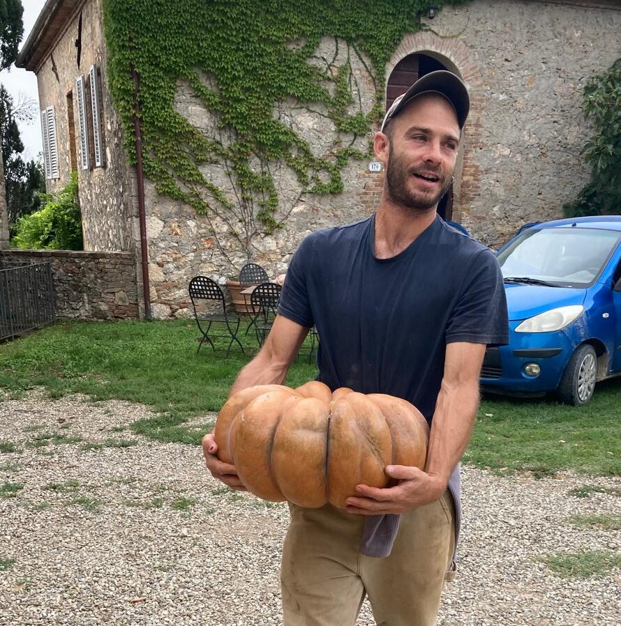 A man, Yago, wearing a baseball hat and blue t-shirt, holding a large orange pumpkin-like gourd. An old stone building and blue car are in the background.