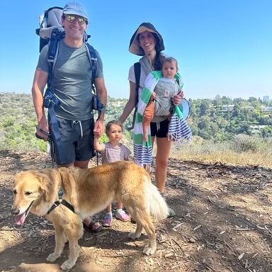 A family: dad, mom, one baby in a front carrier, plus a young girl holding the dad's hand while on a hike. Their golden retriever stands in front of them and there are trees and blue skies in the background.