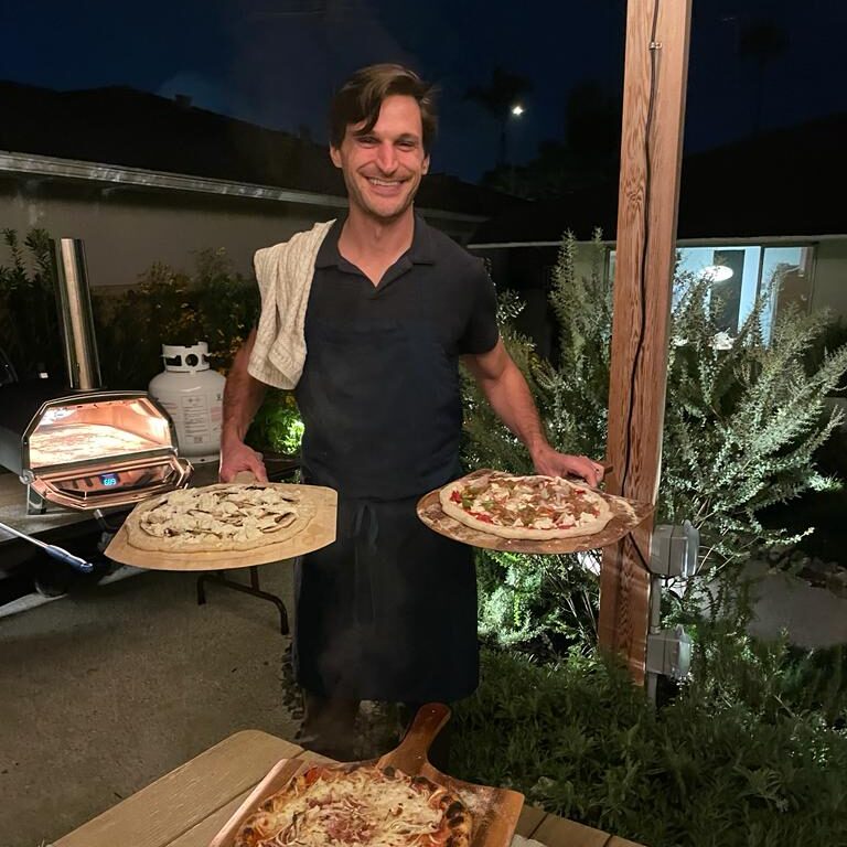 A man, smiling at the camera, in a black polo shirt and apron holding two pizza peels each with a pizza. He is standing outside with some greenery and a pizza oven behind him.