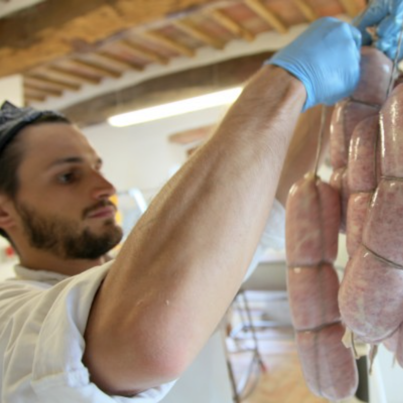 A young man, slightly out of focus on the left of the photo, holding fresh meat tied with twine to be cured, in focus on the right-hand size of the photo. 