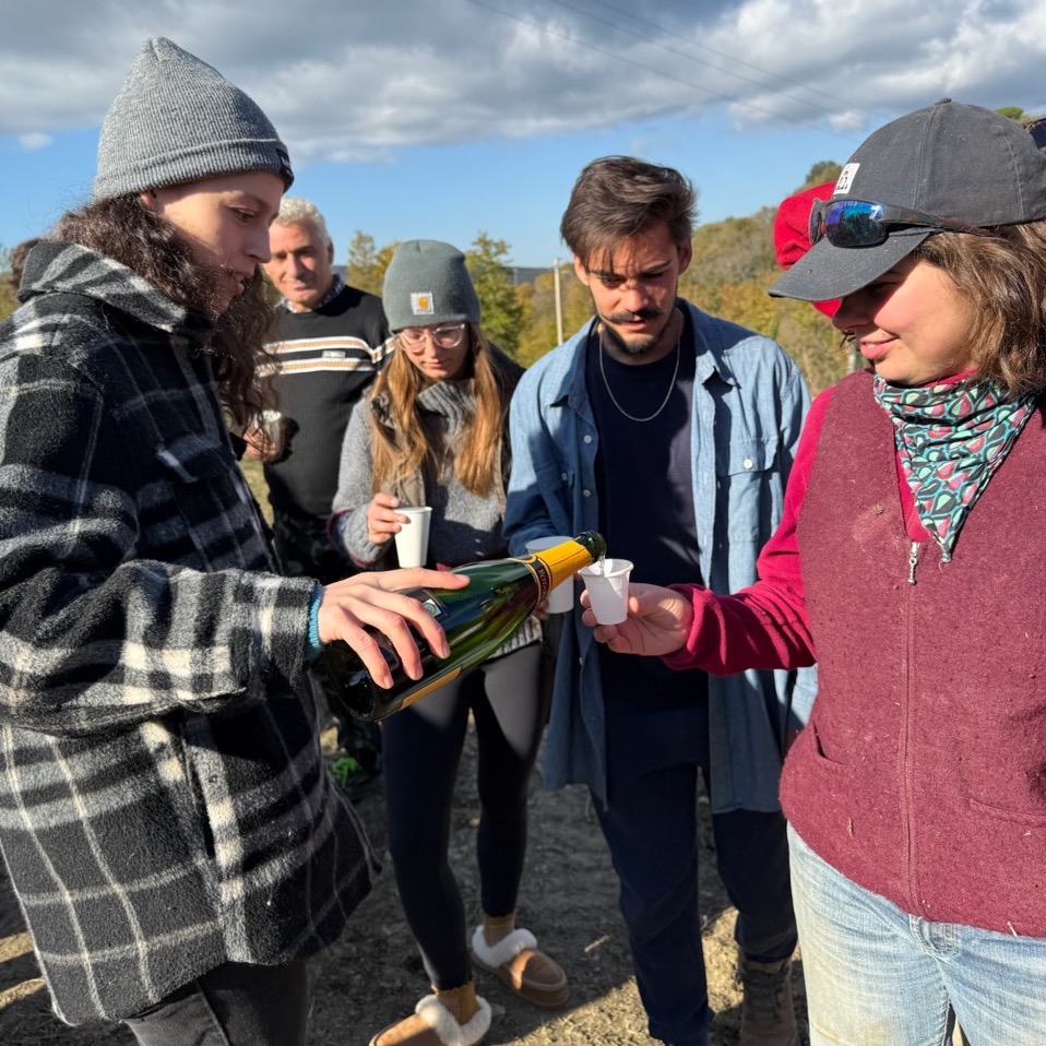 Laura, one of the staff members, pouring sparkling wine into a small cup held by Mary, one of the Fall 2024 interns. Other interns stand beside them.