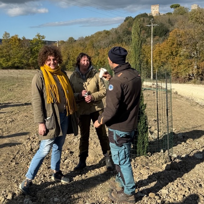 Silvia, Chiara, and Yago stand together next to a cypress tree, talking and sipping sparkling wine. Silvia is smiling at the camera.