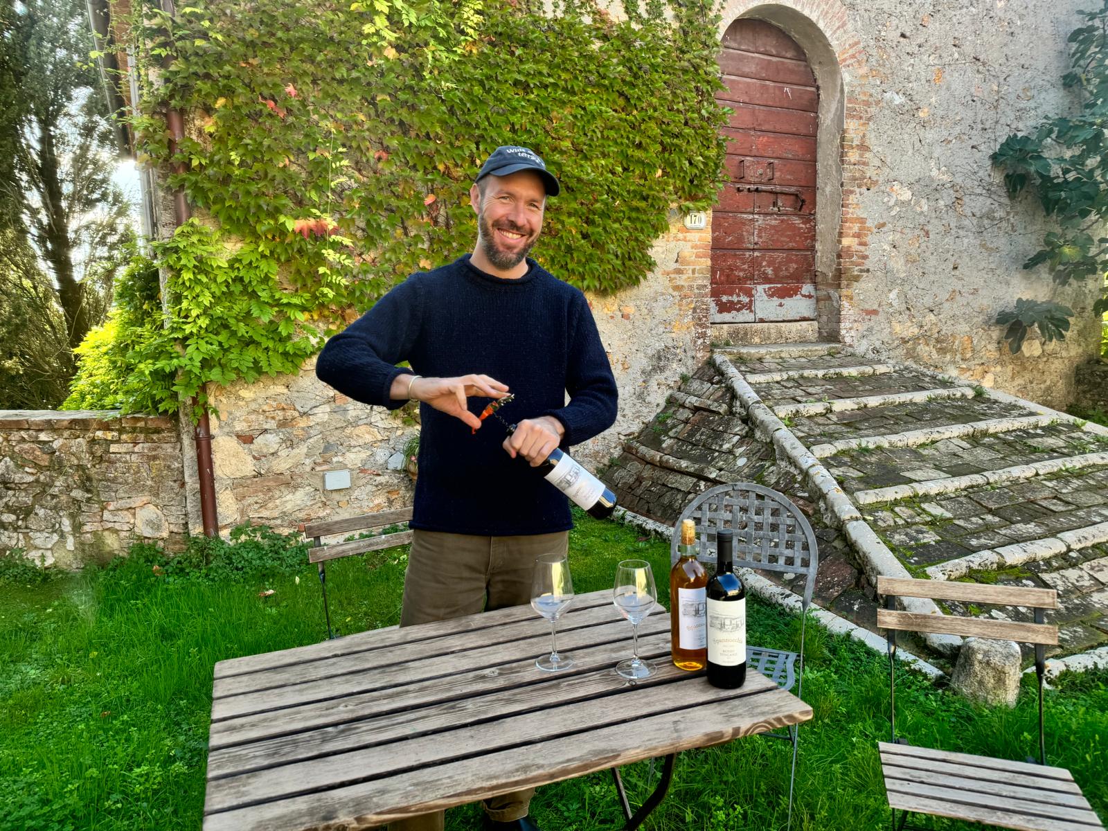 Niccolò stands behind a small wooden table outside, opening a bottle of wine and smiling at the camera. Two more wine bottles and two wine glasses are on the table. An old stone building covered in ivy and very green grass are in the background.