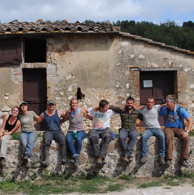 A group of young adults sitting arm in arm on an old stone wall with an old, rustic building behind them.