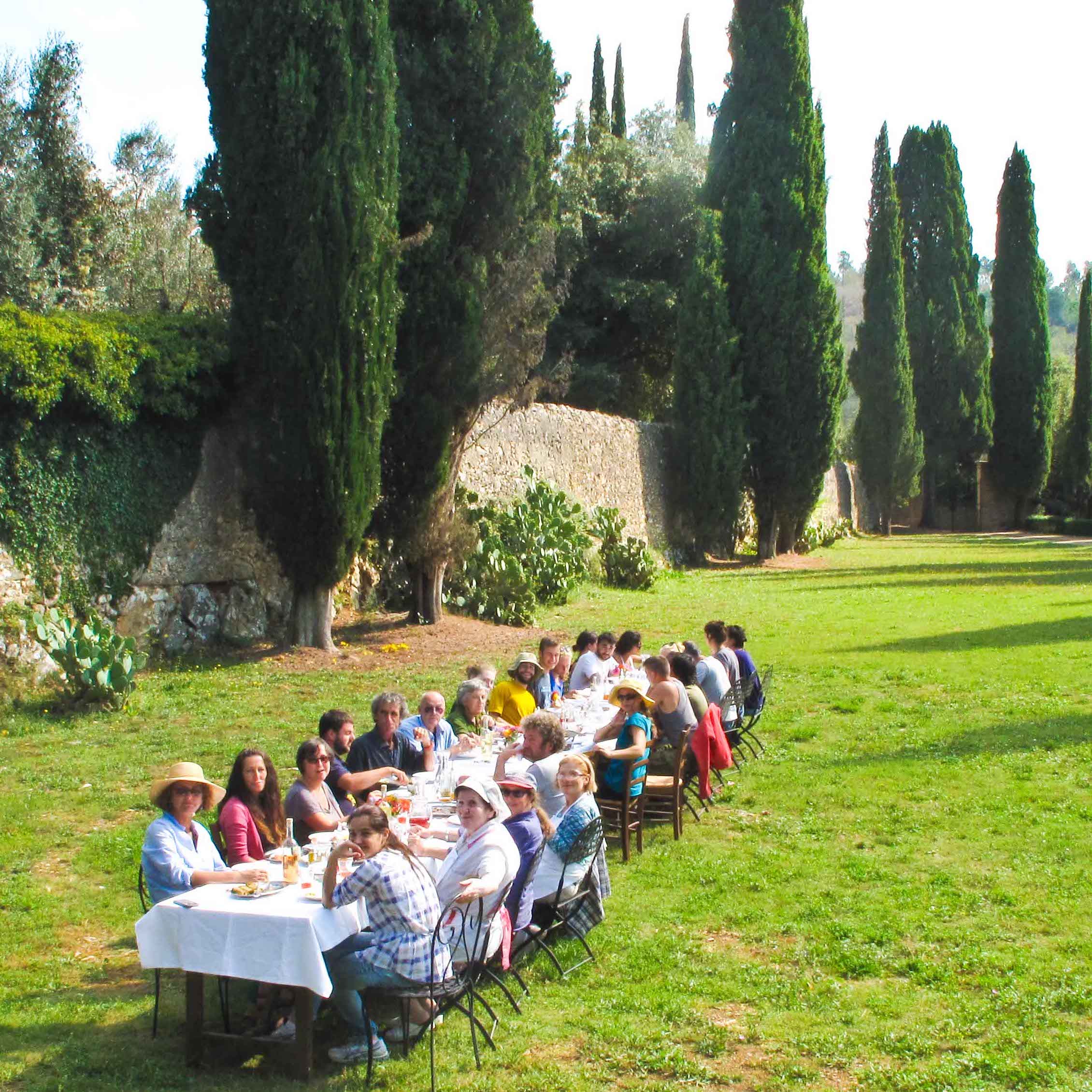 A long table on the grassy Villa lawn, covered in a white tablecloth and surrounded by seated people eating a meal. Cypress trees and an old stone wall are in the background.