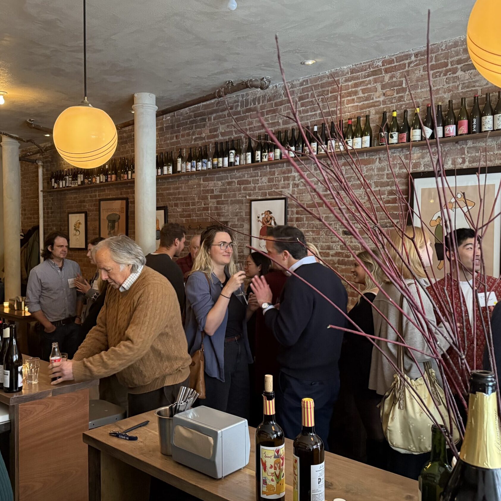 A crowd of people in small groups of conversation in a restaurant space. In the foreground is a bar counter, yellow globe pendants hang from the ceiling, and an exposed brick wall is in the background.