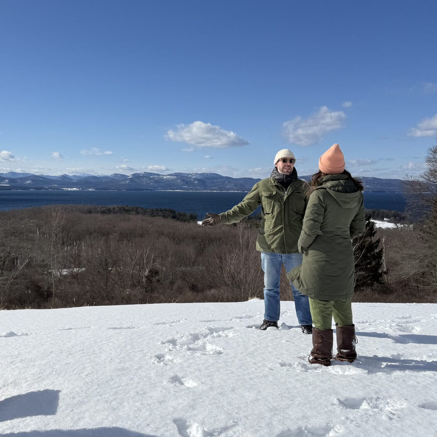 Two people stand in the snow on top of a hill, overlooking a vast expanse of bare trees and then a lake below/behind them. The sky is clear blue with some puffy white clouds.