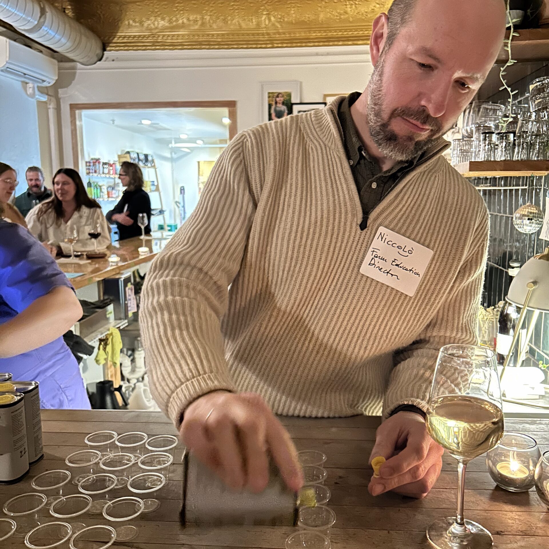 A man, Niccolò, stands behind a butcher block counter in a restaurant space, pouring olive oil from a can into little tasting cups. Some other people are seen in the background.