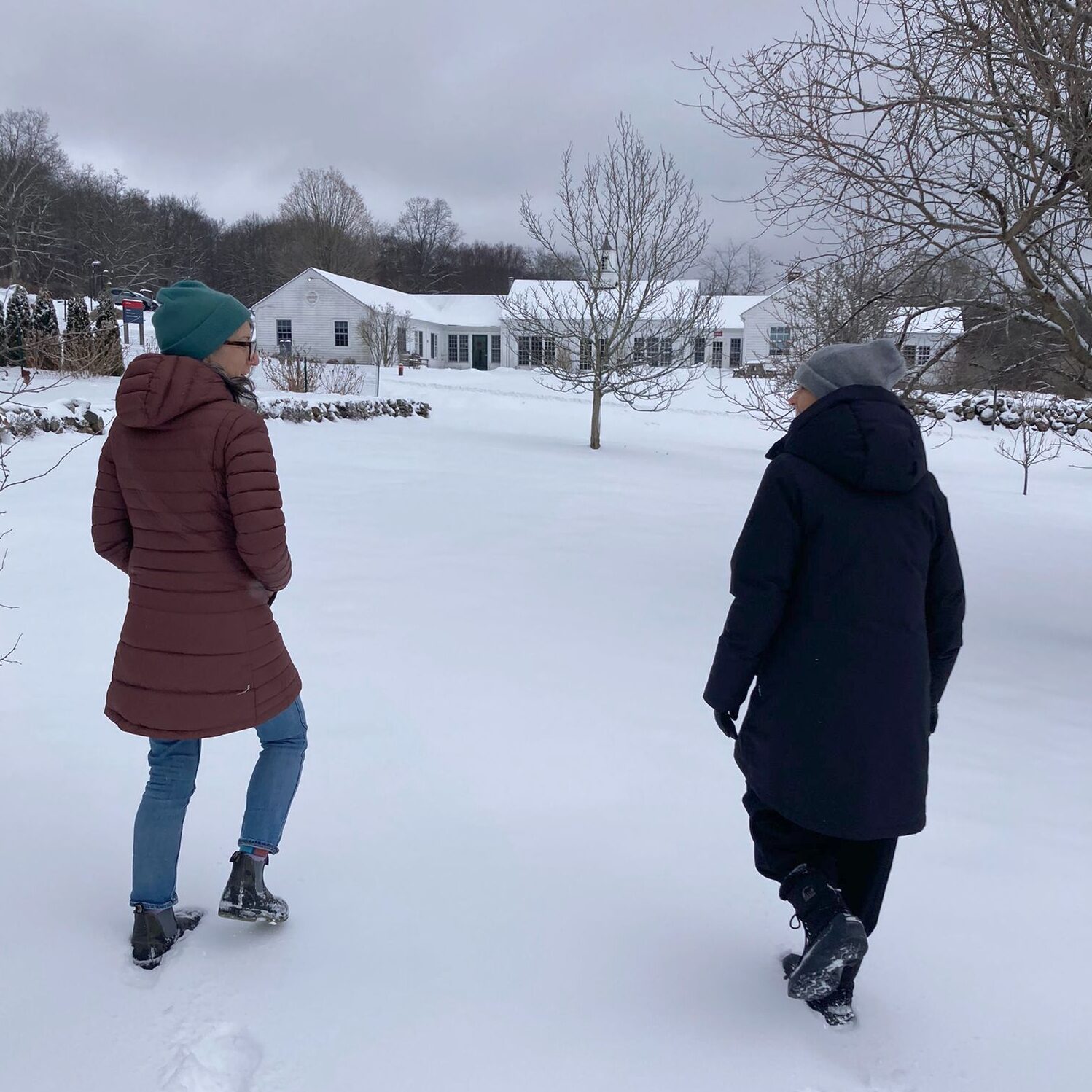 Two people walk with their backs to the camera, bundled up and crossing a snowy field. Bare trees and a low white building are in the background.